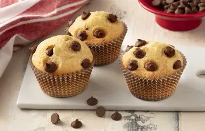 Fotografía en tonos de blanco y rojo de una encimera blanca con un banco, tres muffins encima. Al fondo un paño rojo y un recipiente redondo rojo con chispas de chocolate.