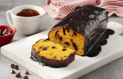 Fotografía en tonos blancos y rojos de una encimera blanca blanca con un plato rectangular blanco, sobre ella un pastel de zanahoria. Al fondo un paño de rayas rojas y blancas y un recipiente redondo blanco con salsa de chocolate.