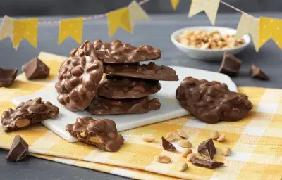 Fotografía en tonos amarillos con un paño a cuadros amarillo y trozos de mocoso chocolate de pie sobre una pizarra blanca. Banderas del partido Junina en cuadros amarillos y lunares en la parte superior y al fondo una pequeña olla blanca con cacahuetes.