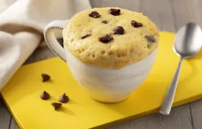 Fotografía en tonos marrones. blanco y amarillo de un banco de madera con un tablero amarillo, sobre él una taza blanca con pastel de taza y una cuchara. Al fondo una tela blanca.