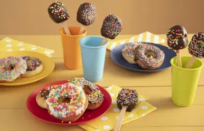 Fotografía en tonos amarillos sobre una encimera de madera amarilla con tres vasos de colores, azul, naranja y verde con piruletas de chocolate en su interior. En el centro, un plato rojo con tres donas cubiertas con chispitas de colores.