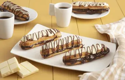 Fotografía en tonos amarillos en un banco de madera con un paño a cuadros color canela, un recipiente rectangular blanco en el centro con tres bombas de chocolate cubiertas de chocolate blanco. Al fondo, dos tazas de café y dos platos con dos bombas.