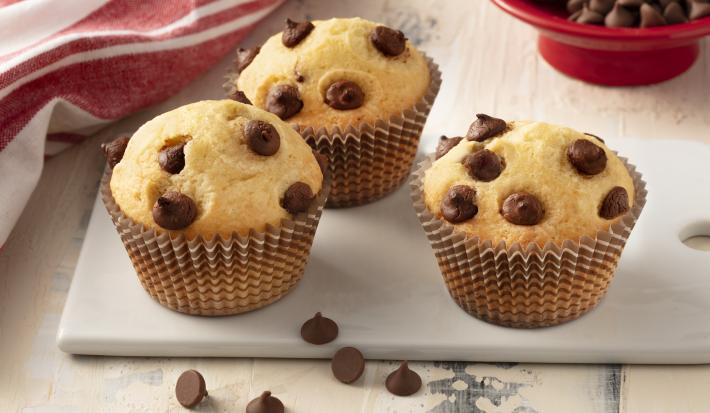 Fotografía en tonos de blanco y rojo de una encimera blanca con un banco, tres muffins encima. Al fondo un paño rojo y un recipiente redondo rojo con chispas de chocolate.