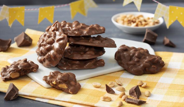 Fotografía en tonos amarillos con un paño a cuadros amarillo y trozos de mocoso chocolate de pie sobre una pizarra blanca. Banderas del partido Junina en cuadros amarillos y lunares en la parte superior y al fondo una pequeña olla blanca con cacahuetes.