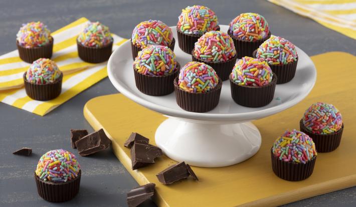 Fotografía en tonos de amarillo sobre una encimera gris oscuro con servilletas amarillas a rayas. En el centro, un tablero amarillo con un soporte de dulces blanco que sostiene los moldes de chocolate con brigadeiros con dulces de colores.