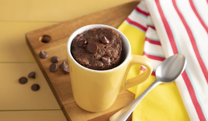 Fotografía en tonos amarillos sobre un banco de madera amarillo, una tela a rayas rojas y amarillas y una tabla de madera en el centro de la foto con una taza amarilla con el pastel de chocolate descansando sobre ella.