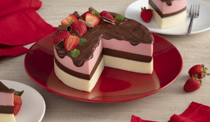 Fotografía en tonos rojos sobre una encimera de madera clara con una placa roja en el centro con la tarta napolitana cortada por la mitad con un glaseado de chocolate cayendo y fresas para decorar. Al lado, paños rojos y fresas esparcidos sobre la mesa.