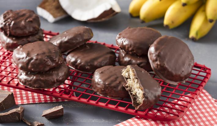 Fotografía en tonos rojos sobre una encimera gris con un respaldo rojo con las galletas de avena, cacao y coco. Al fondo, un racimo de plátanos y trozos de coco.