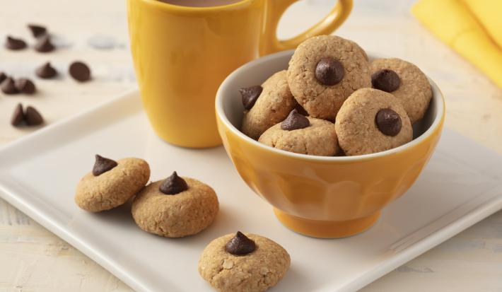 Fotografía en tonos de blanco y amarillo de una encimera blanca con un plato cuadrado, encima una taza amarilla, un recipiente amarillo redondo con galletas y tres galletas. Al fondo un paño amarillo y gotas de chocolate.