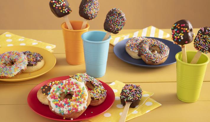 Fotografía en tonos amarillos sobre una encimera de madera amarilla con tres vasos de colores, azul, naranja y verde con piruletas de chocolate en su interior. En el centro, un plato rojo con tres donas cubiertas con chispitas de colores.