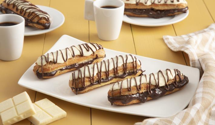 Fotografía en tonos amarillos en un banco de madera con un paño a cuadros color canela, un recipiente rectangular blanco en el centro con tres bombas de chocolate cubiertas de chocolate blanco. Al fondo, dos tazas de café y dos platos con dos bombas.